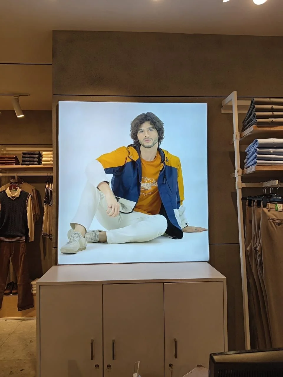 A clothing store display with a large framed photograph of a young man with curly hair, wearing a yellow jacket over a blue vest and white pants, sitting on the floor against a white background.