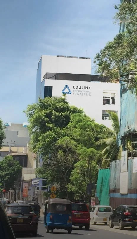 Street view with cars and trees, a large white building in the background with a sign that reads 'Edulink International Campus'.