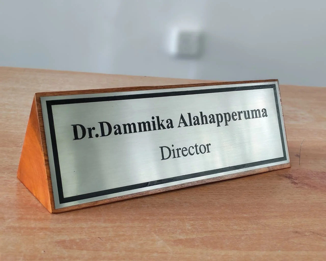 A nameplate on a wooden table reading 'Dr. Dammika Alahapperuma, Director' with a blurred white wall in the background.