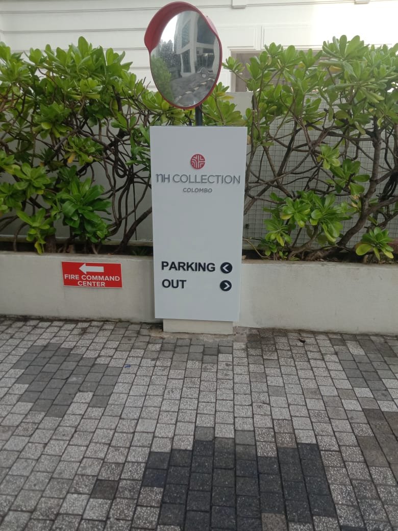 Sign for NH Collection Colombo hotel indicating parking directions, with a red fire command center sign to the left and lush green shrubs in the background.
