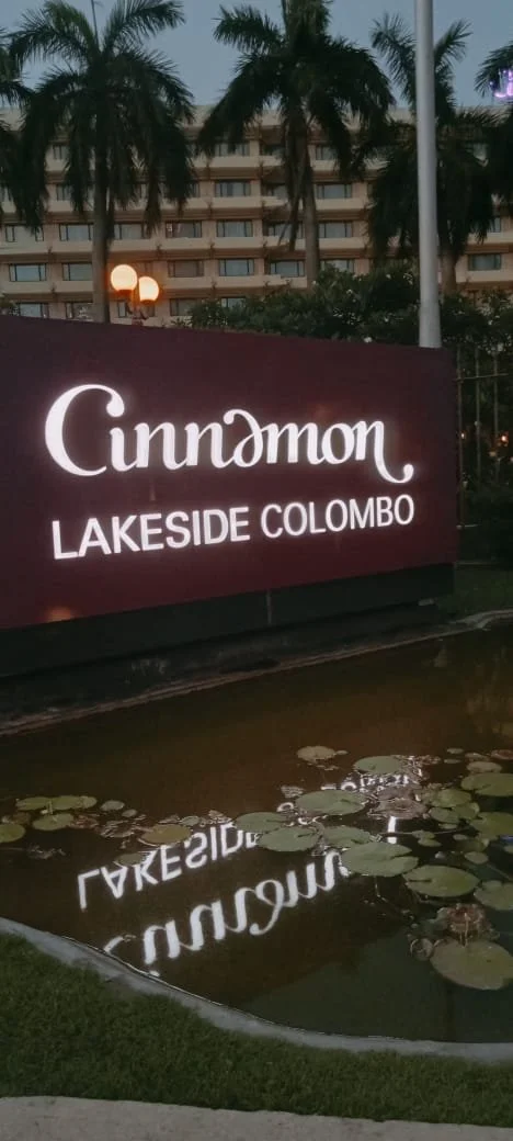 A sign with the text "Cinnamon Lakeside Colombo" near a pond with floating water lilies, with palm trees and a multi-story building in the background.