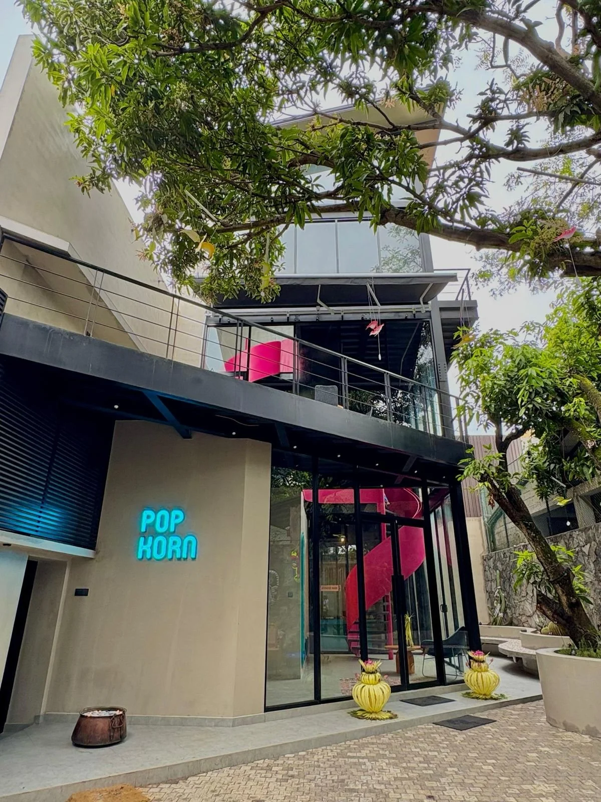 Modern building with a glass entrance, a neon sign that reads "POP KORN", pink slide visible inside, and greenery including trees and hanging flowers outside.