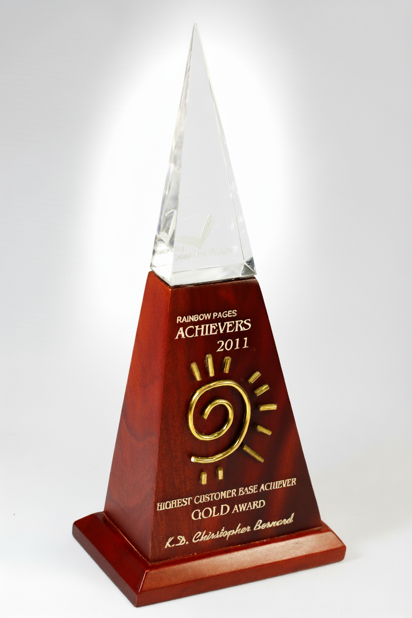 Triangular award with a wooden base and a clear glass top, engraved with the text 'Rainbow Pages Achievers 2011 Highest Customer Base Achiever Gold Award K.D. Christopher Bennett' and a gold spiral symbol.