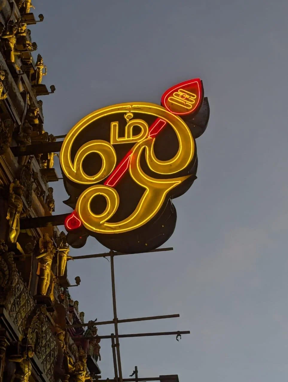 Neon sign with a yellow stylized Hindu goddess Ganesha's face, red trunk, and red ear, mounted on a building with detailed gold ornamentation, against a dark evening sky.