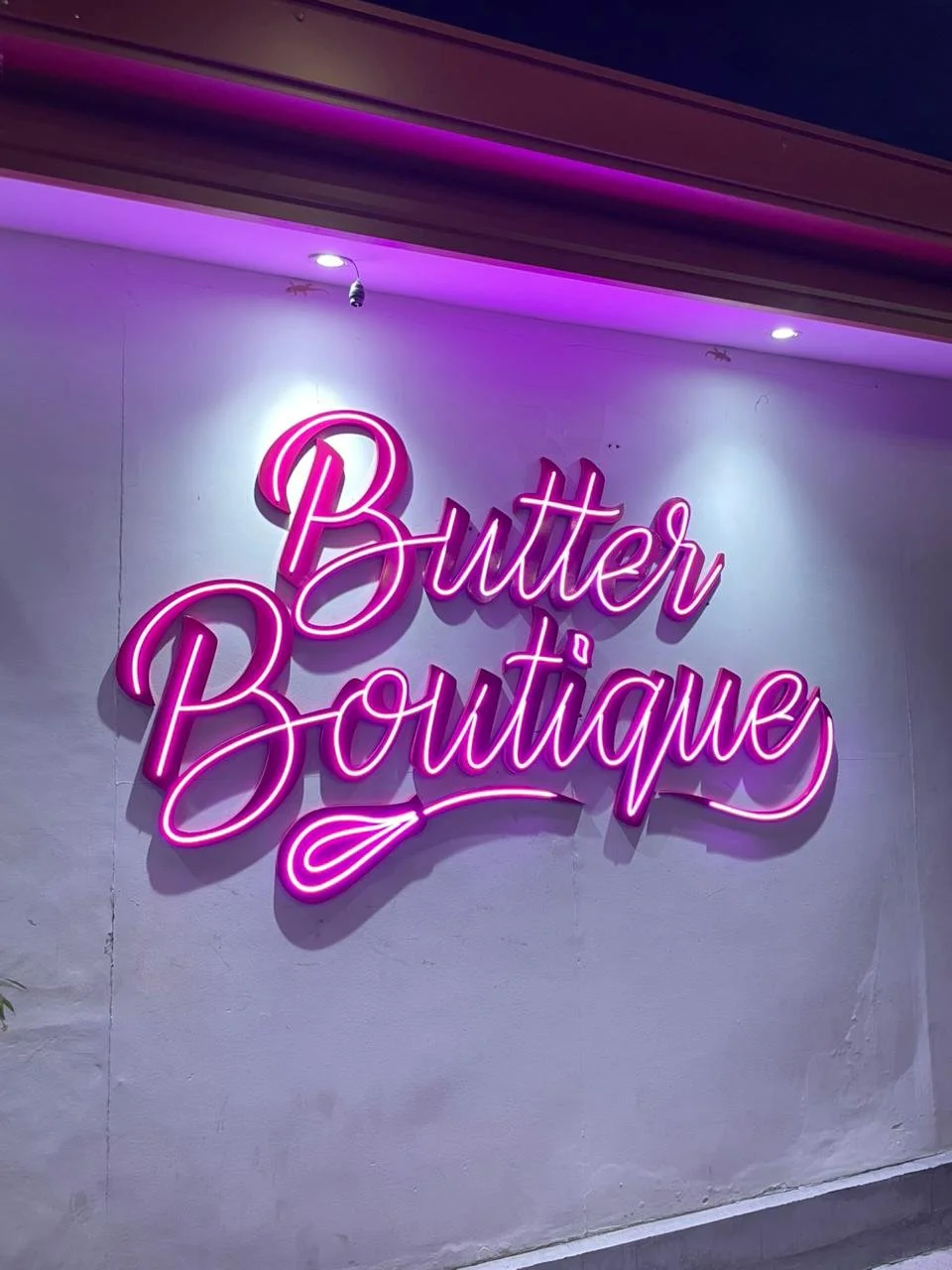 Neon sign reading 'Butter Boutique' in pink cursive letters on a white wall with purple accent lighting.