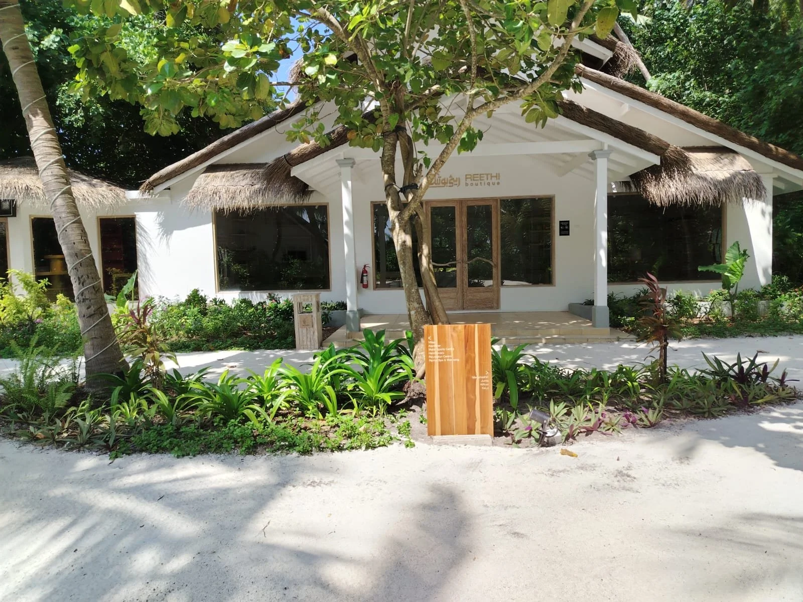 A white boutique building with thatched roofs and large windows, surrounded by lush green plants and trees.
