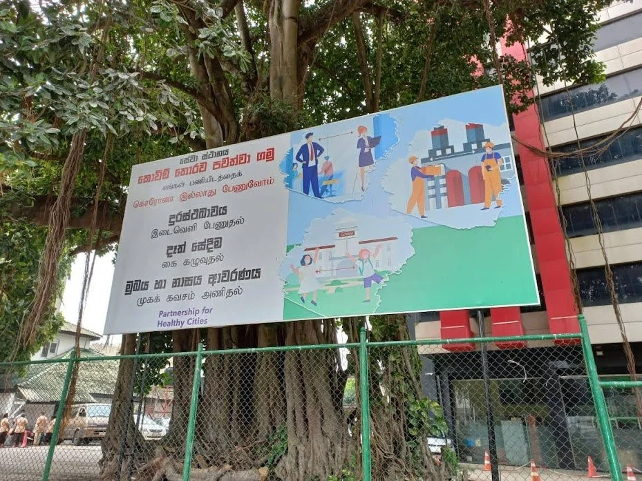 A large billboard sign hanging on a tree, with illustrations and text in a language other than English, promoting partnership for healthy cities. The illustrations depict people working together, urban infrastructure, and environmental activities.