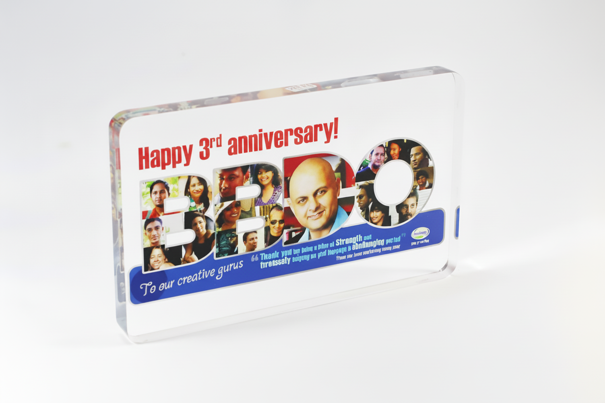 A clear, rectangular acrylic plaque with the text "Happy 3rd anniversary!" in red above a collage of photos inside the number 30. There is a message in blue that reads "To our creative gurus" and a brief paragraph underneath.