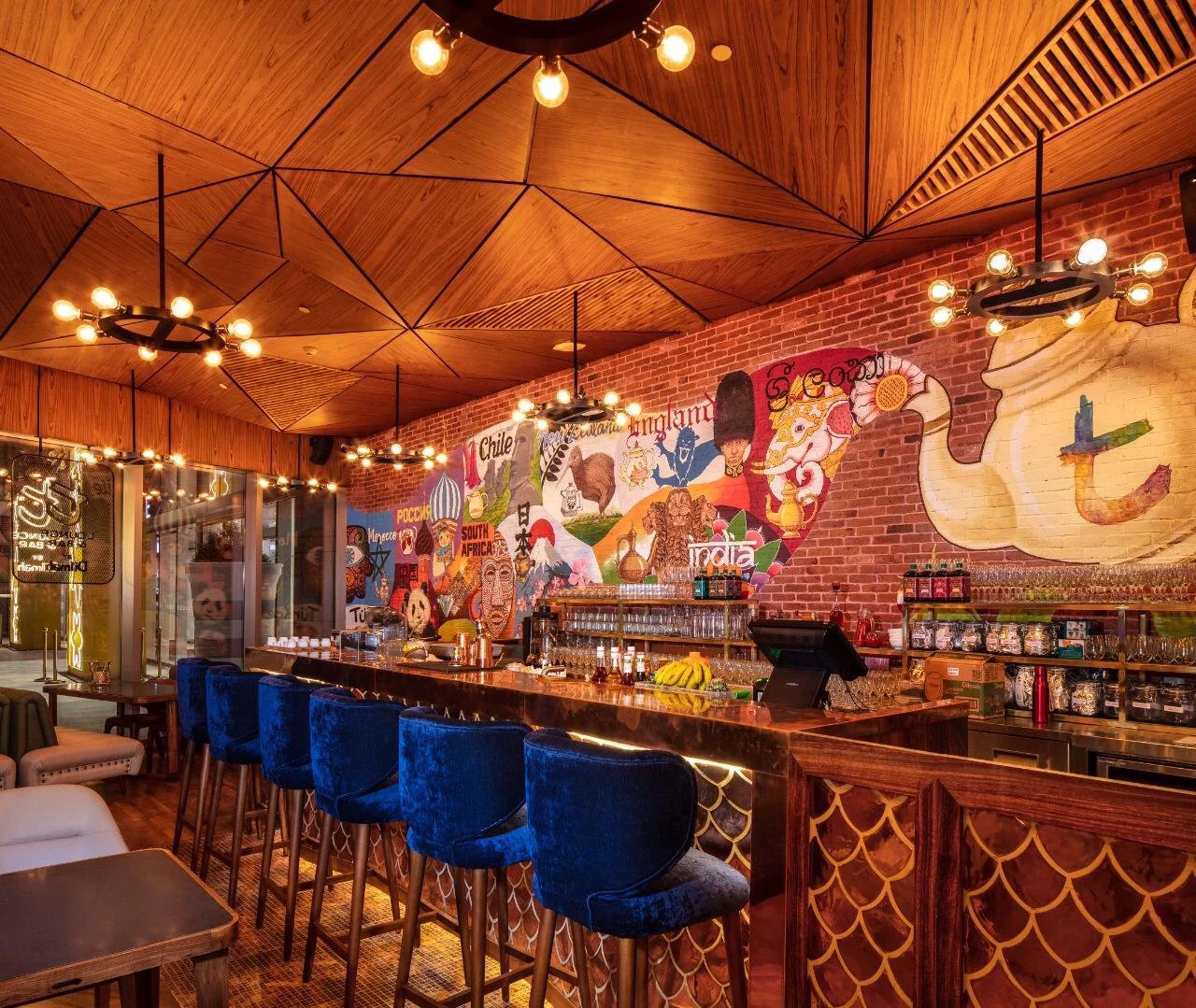Interior of a restaurant or bar with a mural of world flags and symbols on a brick wall, wooden ceiling with geometric patterns, and a bar counter with blue velvet chairs.