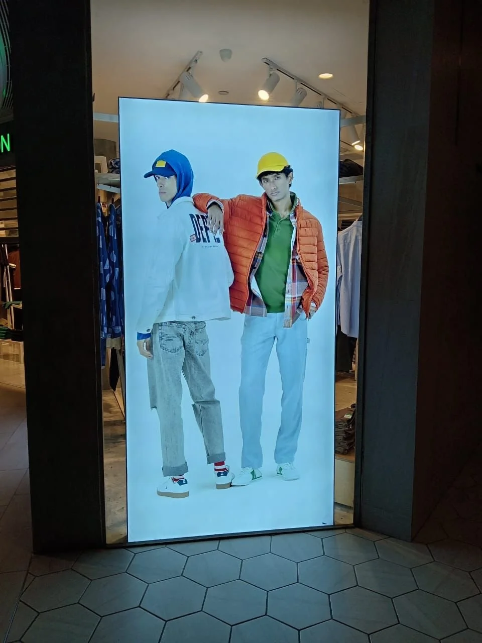 Store advertisement featuring two young male models dressed in casual, sporty clothing, with one wearing a blue cap and hoodie and the other wearing a yellow helmet and orange jacket.