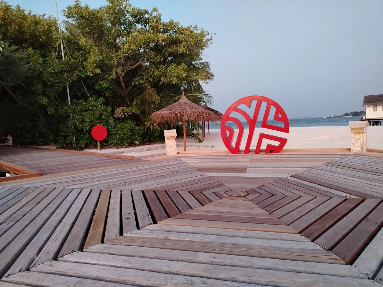 Empty wooden boardwalk on a beach with greenery, a thatched umbrella, a red decorative circle, and a circular red and white abstract sculpture by the water.