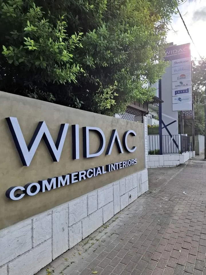 Sign for WIDAC Commercial Interiors with large 3D letters on a concrete wall and a tall pole sign with various business logos, near a sidewalk with greenery in the background.