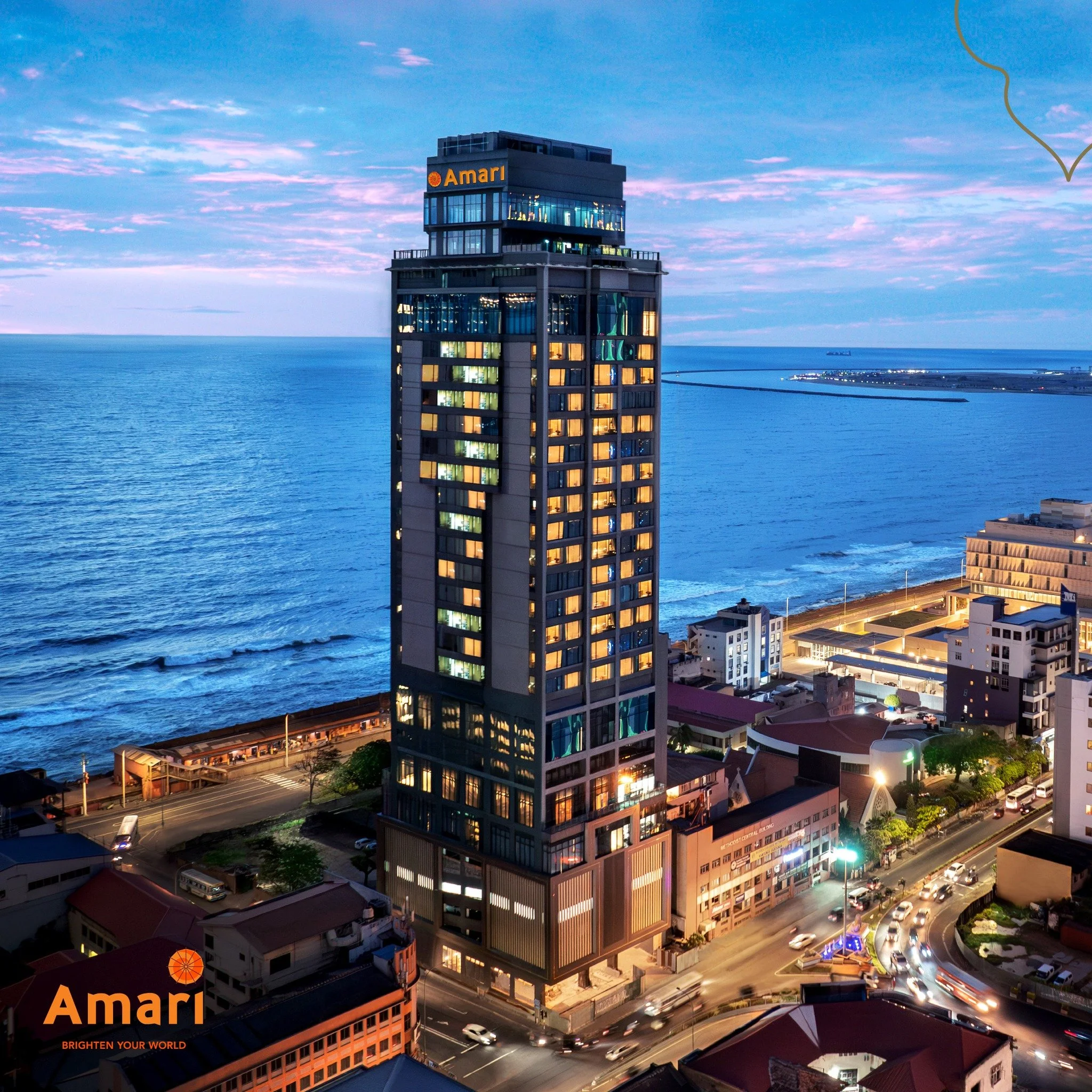 A tall modern building with the logo 'Amari' on top, overlooking a body of water. The building has illuminated windows and is located in a city with busy streets and cars. The sky is at dusk with scattered clouds.