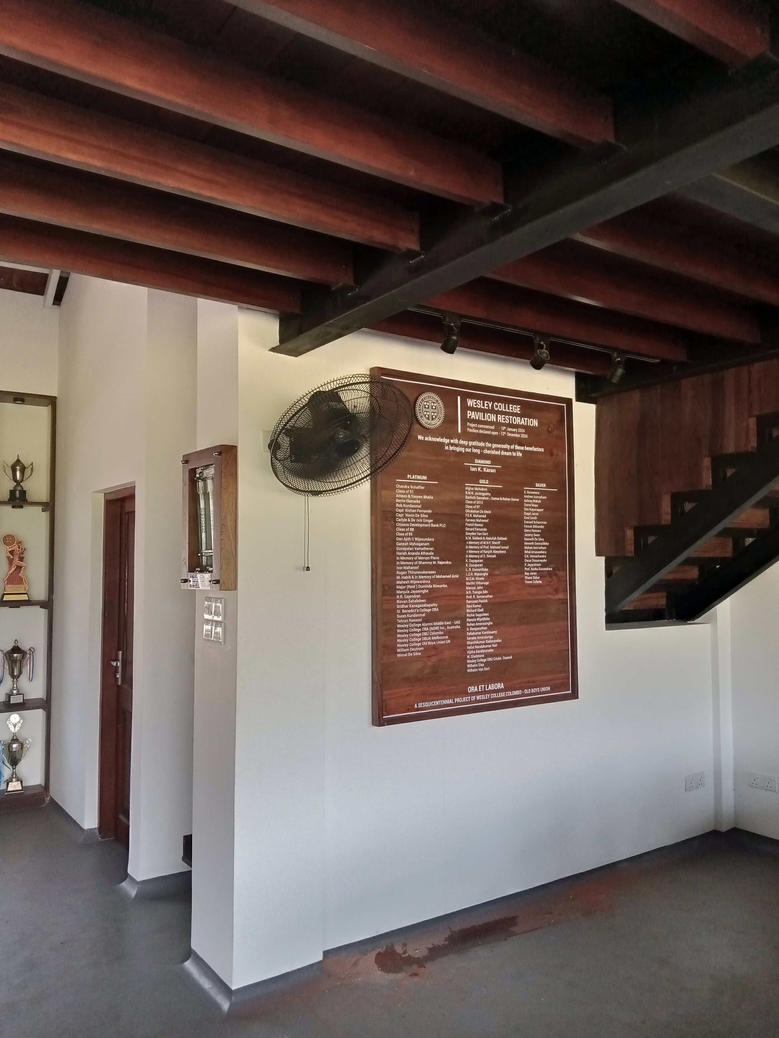 Interior of a building with a staircase, a fan, a wooden plaque with names, and a display shelf with trophies.