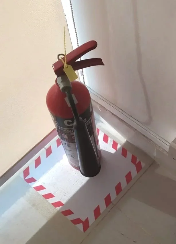 A fire extinguisher turned upside down with a tube hanging from it, placed on a red and white striped cardboard tray on the floor near a wall.