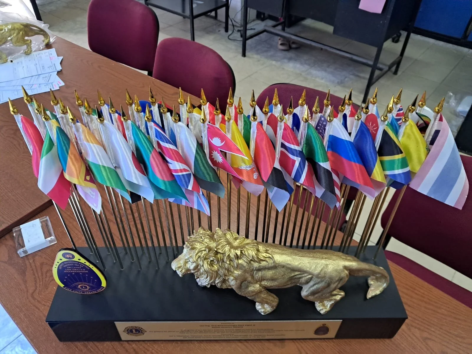 A display of small international flags on gold poles, with a gold lion sculpture at the front, on a table likely for an event or organization.