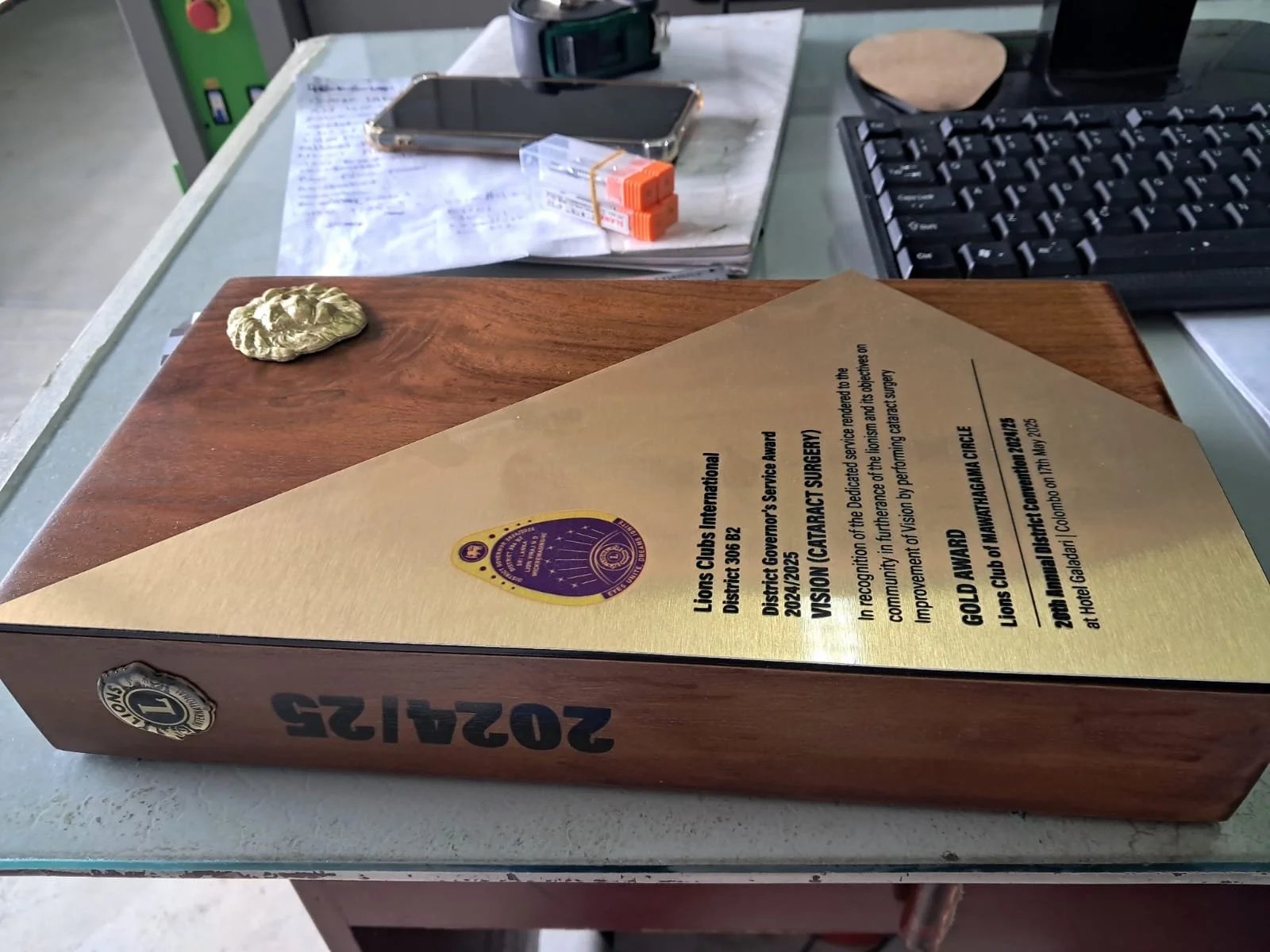 A desk with various items including a certificate, a smartphone, sticky notes, a keyboard, and a wooden board with a gold-colored decorative object attached.