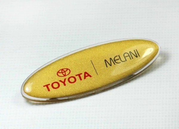 A gold-colored keychain with the Toyota logo, the word 'TOYOTA' in red, and the names 'MELANI' separated by a vertical line.