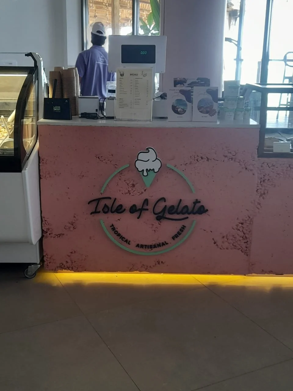 Pink ice cream counter with 'Isle of Gelato' logo, showing a small ice cream cone icon with a scoop, and the words 'Tropical Artisanal Fresh'. A worker in a purple shirt and white cap stands behind the counter, with a menu and ice cream display visib
