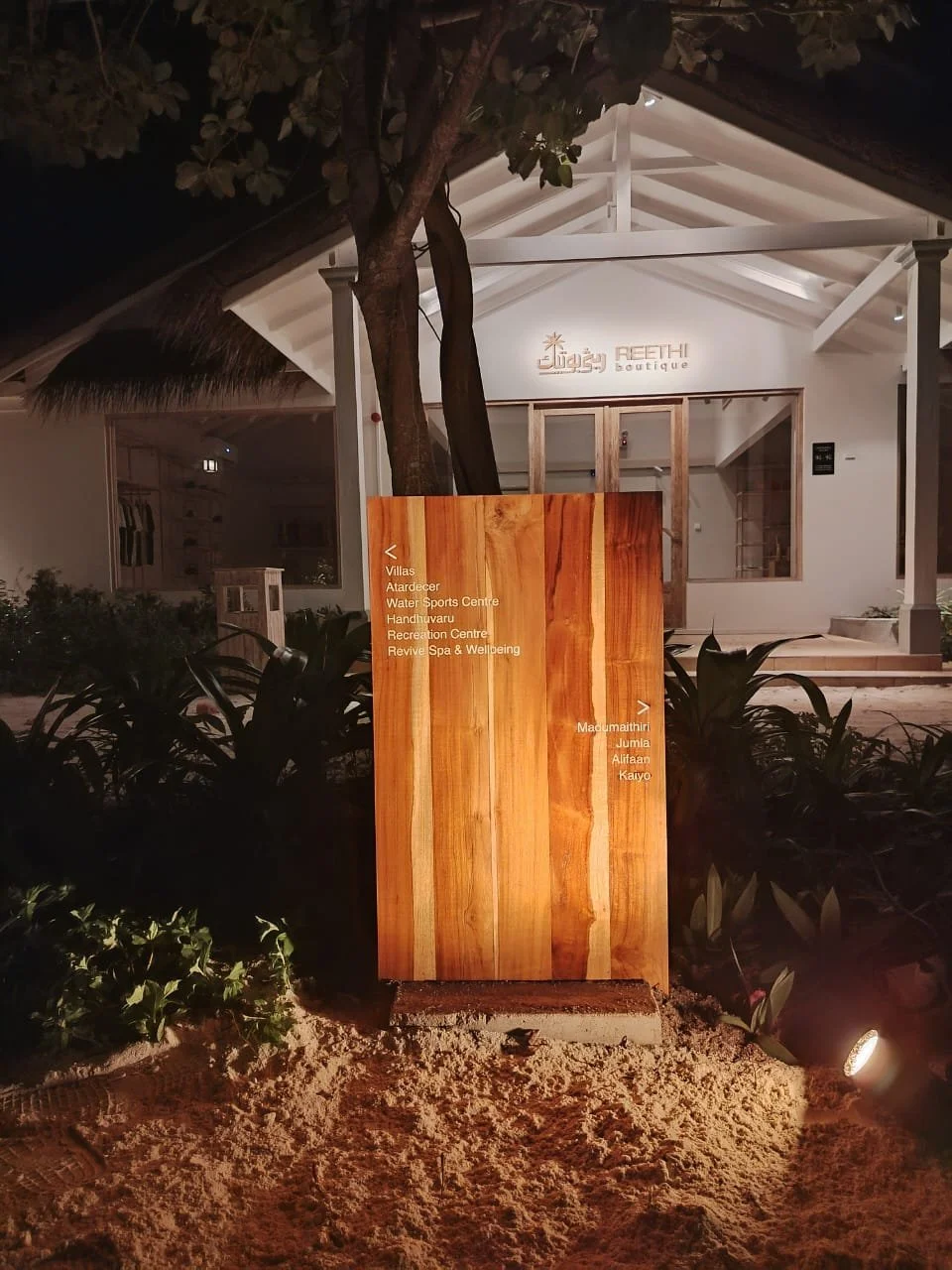 Nighttime view of a white building with a sign that reads 'Reethi Boutique' behind a wooden directional sign with white text, surrounded by plants and illuminated by ground lights.
