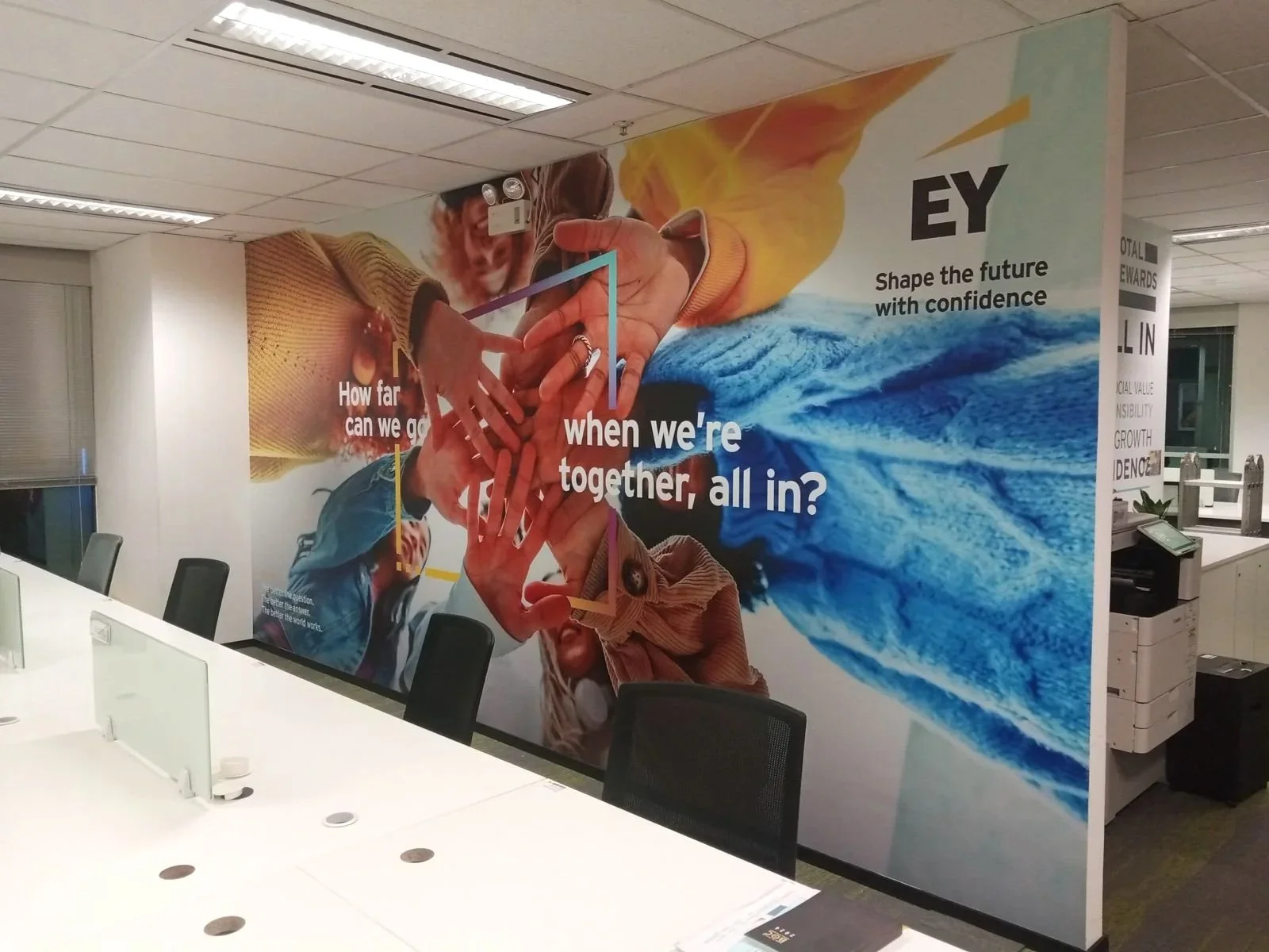 Corporate office conference room showing a large mural on the wall with hands reaching toward each other, water, and colorful abstract elements. Part of the mural reads, 'How far can we go when we're together, all in?' and 'Shape the future with confidence'.