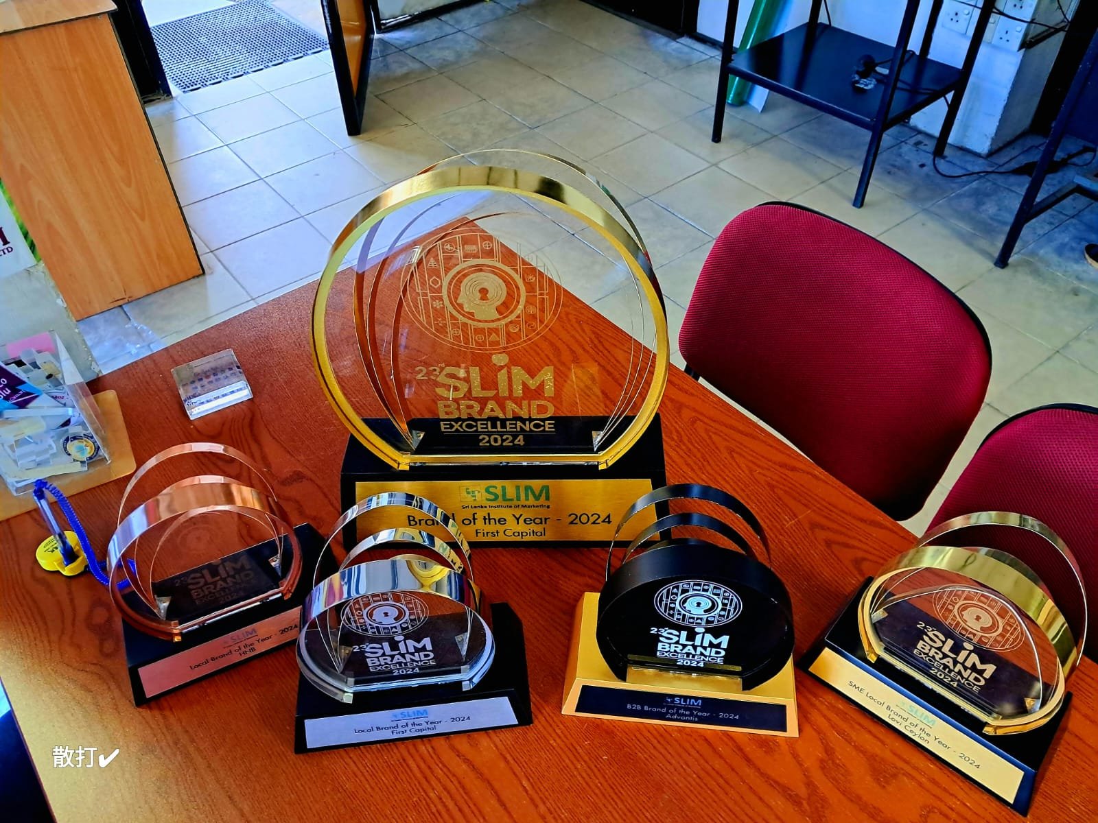 A collection of awards and trophies on a wooden table, including a large acrylic trophy with gold and black accents that reads '23rd SLIM Brand Excellence 2024' and smaller similar awards with labels such as 'Local Brand of the Year 2024 First Capita