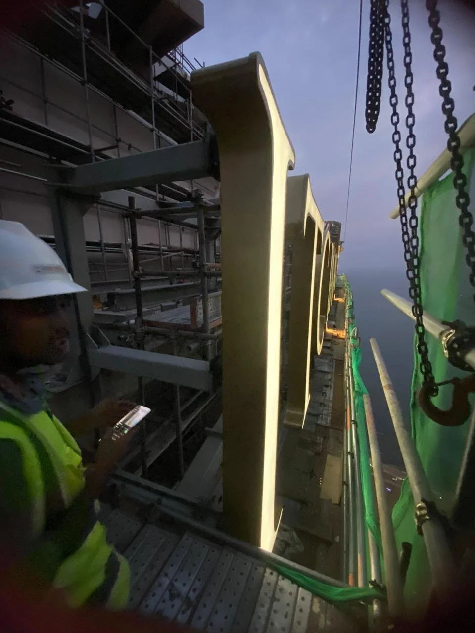 Construction workers in safety gear working on a large outdoor sign installation at dusk, with scaffolding and green safety barriers.