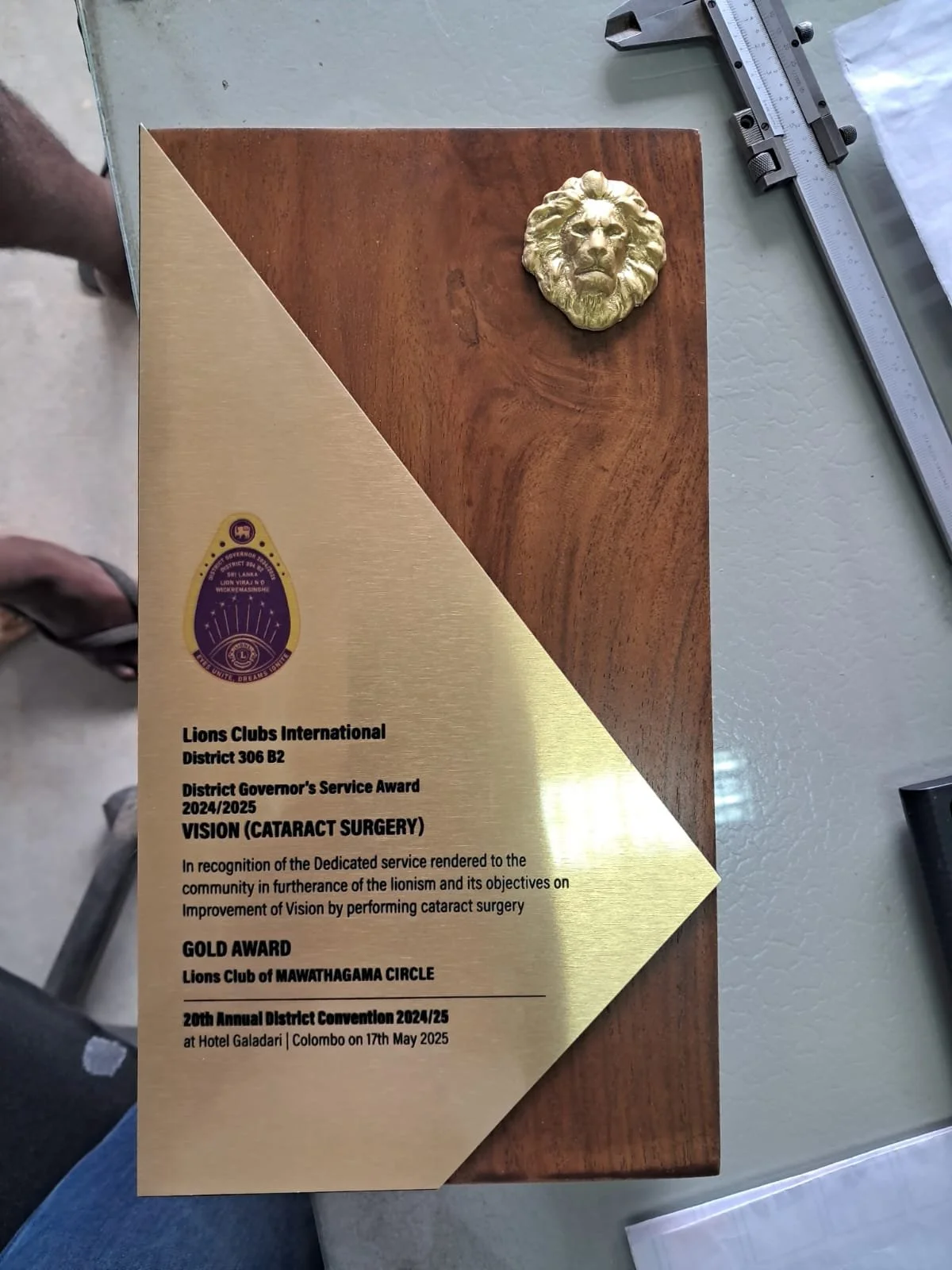 A certificate or award plaque from Lions Clubs International recognizing a service achievement in cataract surgery. The plaque has a gold triangular design with an emblem at the top left corner, and text detailing the award and event, including the L