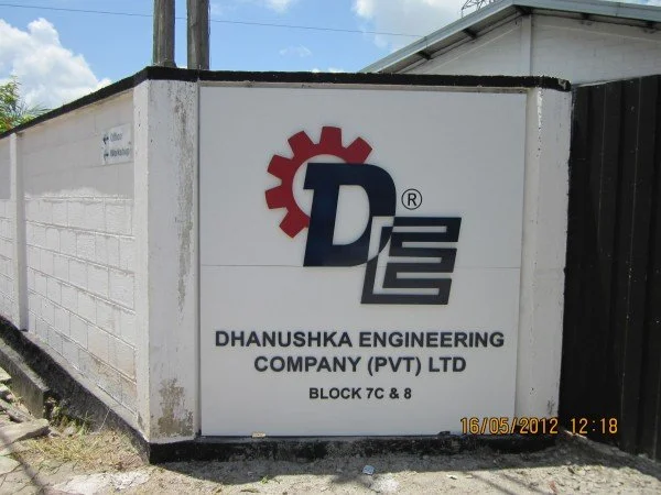 Sign for Dhanushka Engineering Company (PVT) LTD on a white wall, featuring a red gear icon and the company initials D E, with location details.