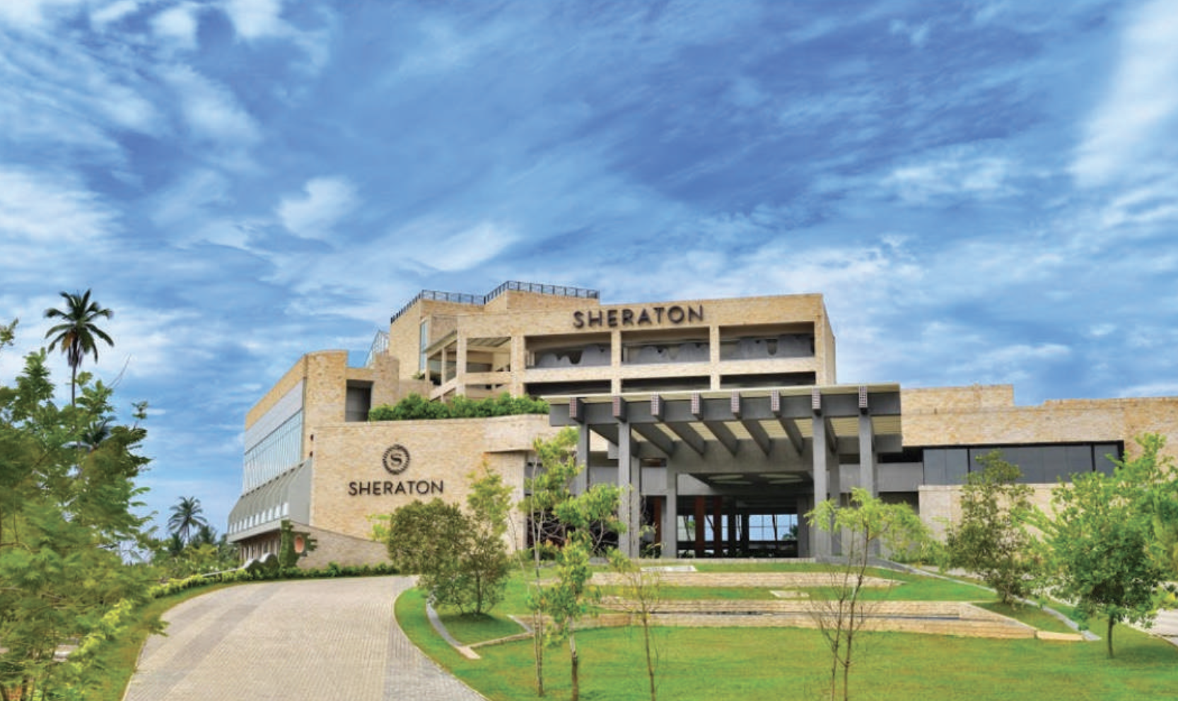 Sheraton hotel building with a landscaped driveway, surrounded by green trees, under a partly cloudy sky.