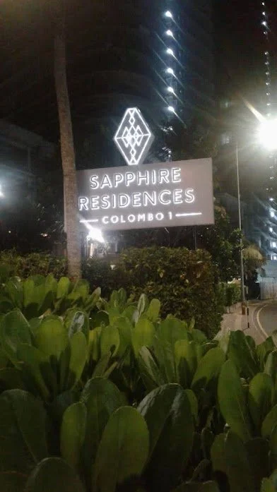 Nighttime view of a sign that reads 'Sapphire Residences Colombo' with a geometric logo above, surrounded by greenery and tall buildings in the background.