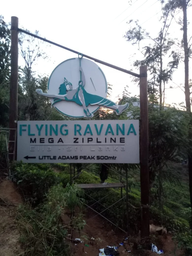 Sign advertising the Flying Ravana Mega Zipline near Little Adams Peak, with a colorful cartoon illustration of a person flying with a Ravana illustration, set in a lush, wooded area.