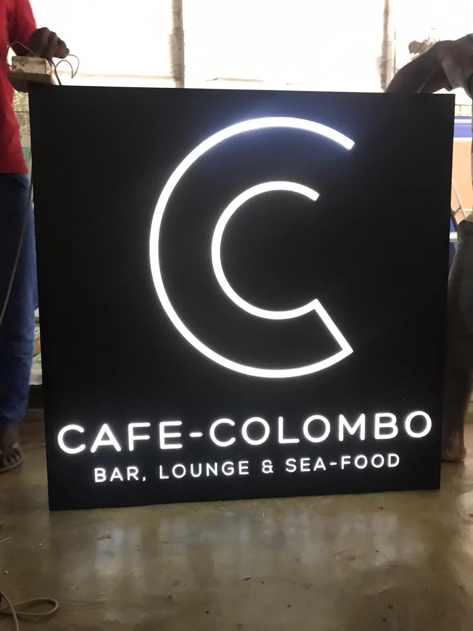 Illuminated black sign for Cafe Colombo with a large letter C and the text 'Bar, Lounge & Sea-Food' below.
