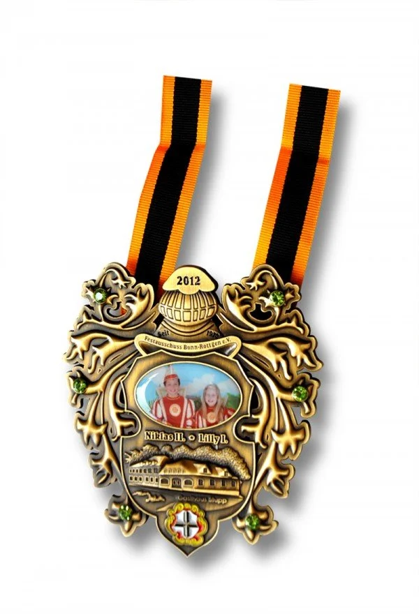 A decorative medal with a ribbon, featuring a photo of two children in historical costumes, a train, and various symbols and text, including "2012" and the names Nickel and Lilly.