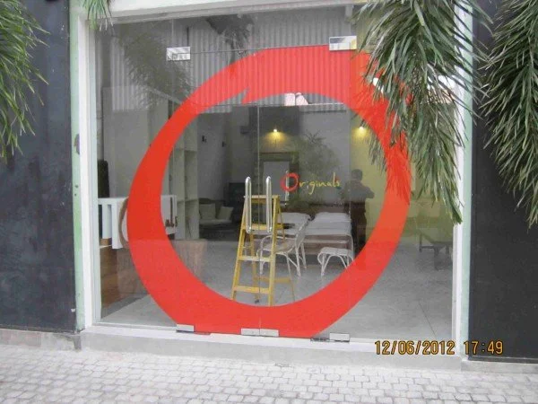A storefront window with a large red circle painted on the glass. Inside, there are chairs, a ladder, a sofa, and a neon sign that says 'Originals.' Potted plants are outside on either side of the window. The photo has a timestamp of December 6, 2012