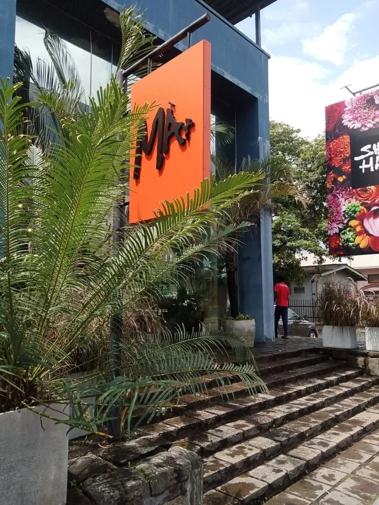 Exterior of a building with large glass windows, surrounded by palm plants and greenery, featuring a bright orange sign with black cursive text.