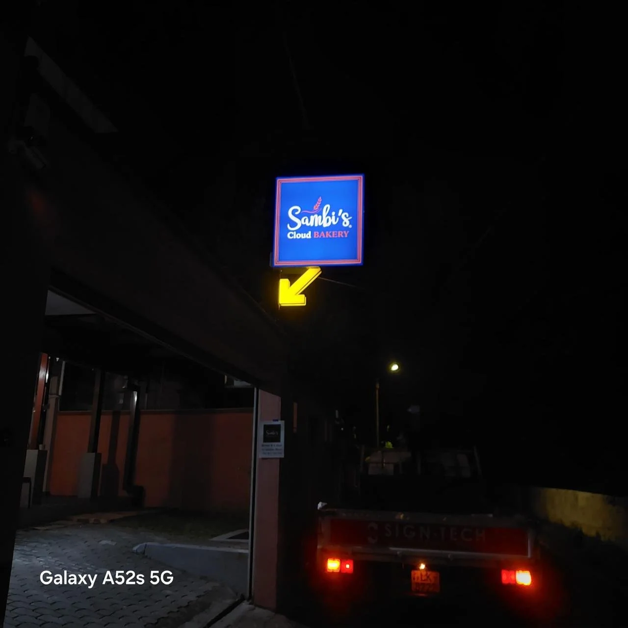 Neon sign for Sambi's Cloud Bakery with a yellow arrow pointing left, visible in a dark outdoor setting at night.