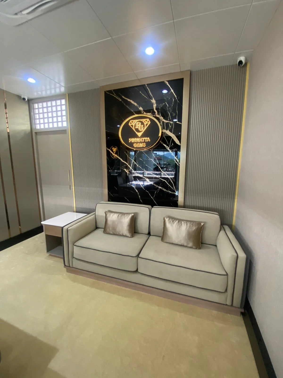 A modern lounge with a beige sofa, two metallic pillows, a small white side table, a black marble wall with gold veins, and a neon sign reading 'Pinwatta Gems' with a diamond logo.