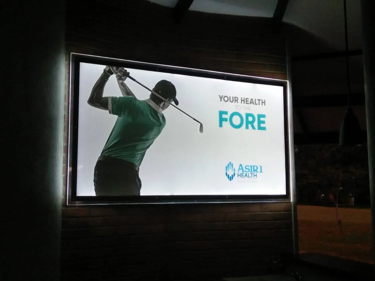 Outdoor illuminated advertisement featuring a man in a green shirt and black cap preparing to swing a golf club, with text promoting health and fitness by ASIRI Health.