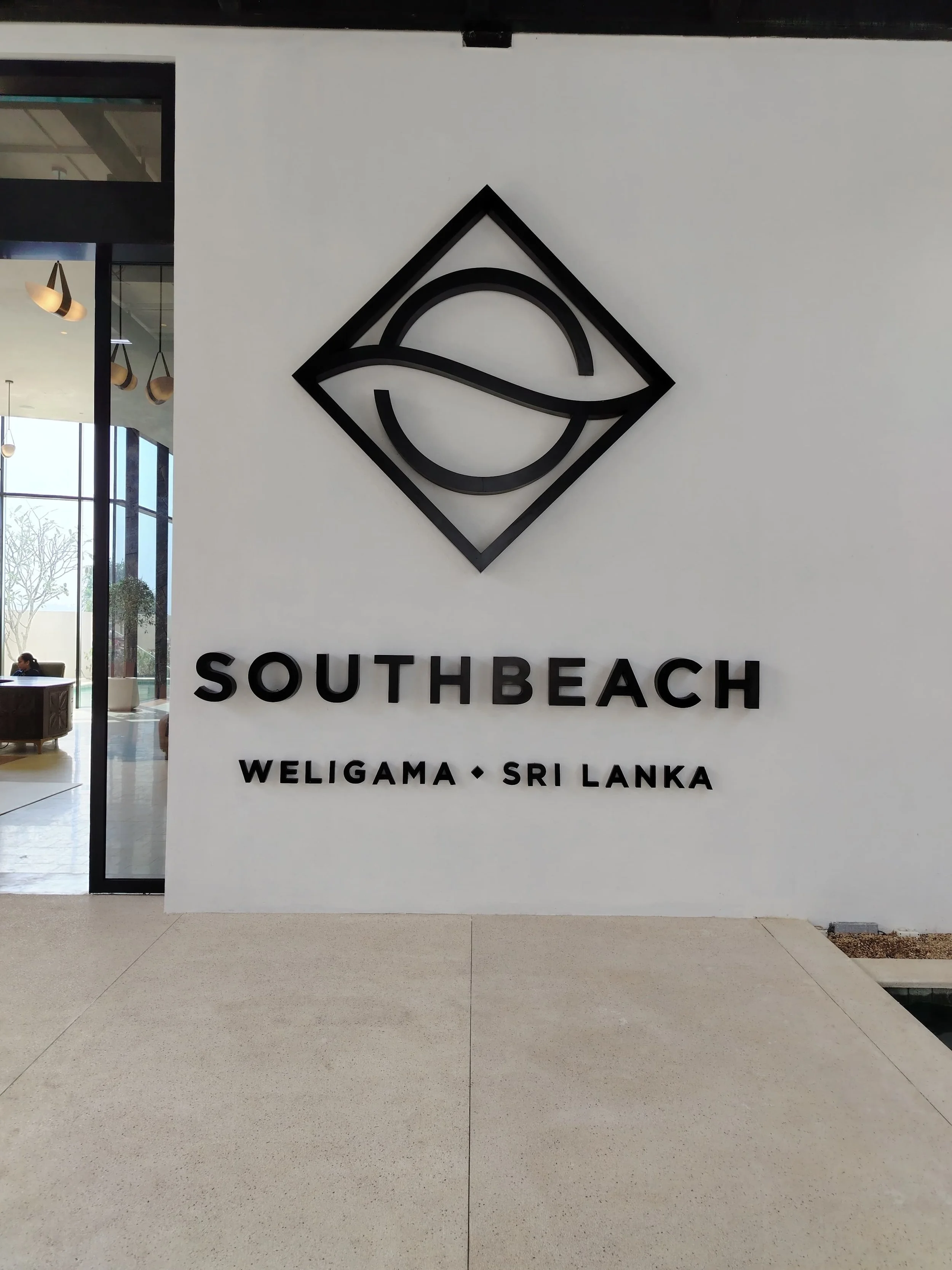 Sign with the words "SouthBeach" and "Weligama - Sri Lanka" underneath, along with a black abstract logo featuring geometric shapes on a white wall.