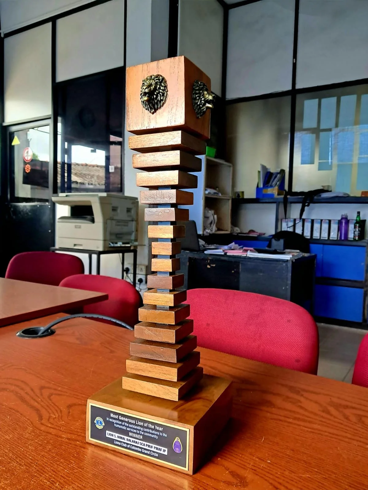 A trophy with a chessboard pattern made of wooden blocks, topped with two lion head ornaments, displayed on a table in an office setting. The trophy has a plaque recognizing the Lions Club of Colombo Grand Circle for their contributions.
