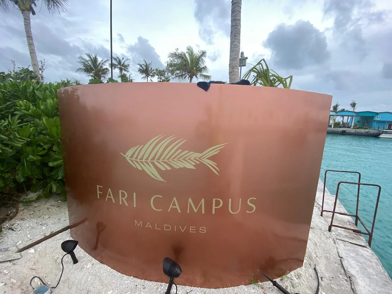 Large brown sign with a feather fish logo and the text 'Fari Campus Maldives' near the water with palm trees in the background.