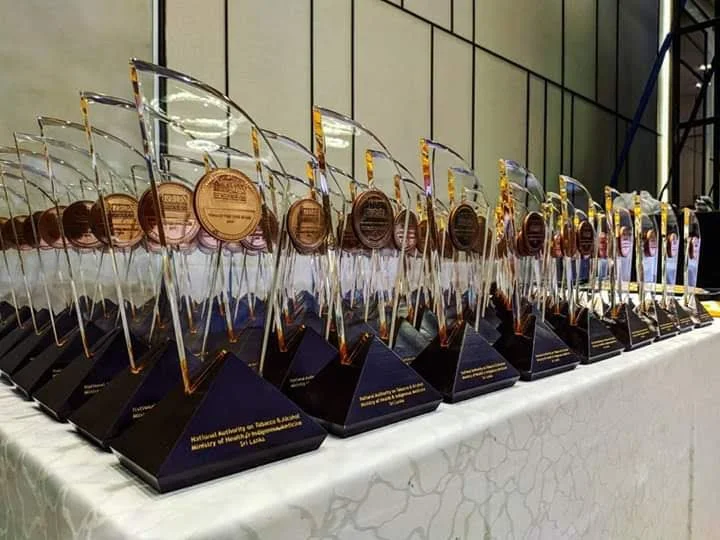 A table lined with multiple awards featuring gold medals and dark triangular bases, set against a backdrop of a large glass window.