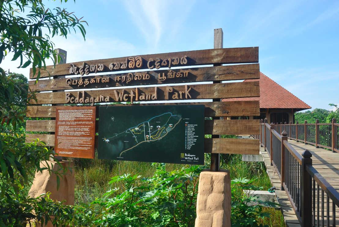 Signboard for Beddagana Wetland Park with a map and informational text, a wooden walkway, and greenery in the background.