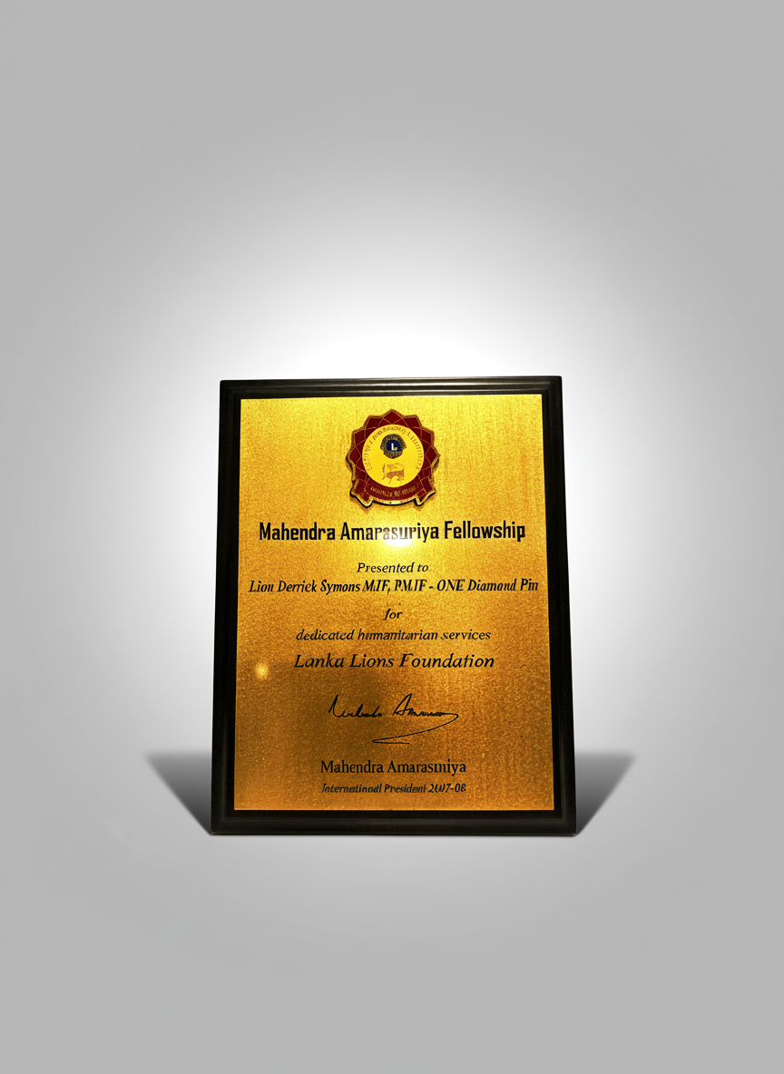A framed award plaque with a gold background, presented to Lion Derniek Symons from Mahendra Amarasuriya Fellowship by the Lanka Lions Foundation, signed by Mahendra Amarasuriya, with the Lions emblem at the top.