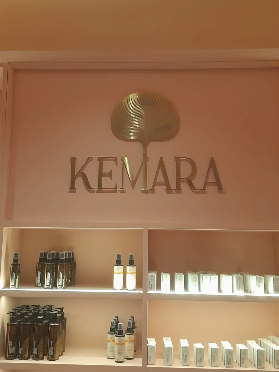 Store display with shelves of skincare products in front of a pink wall, with a metallic logo that says 'KEMARA' and features a stylized gold leaf above the name.