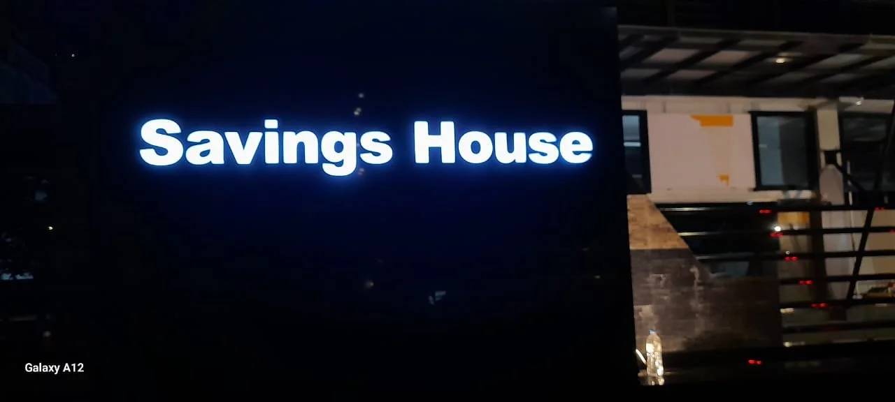 Illuminated sign reading 'Savings House' against a dark background, with part of a building structure visible in the background.