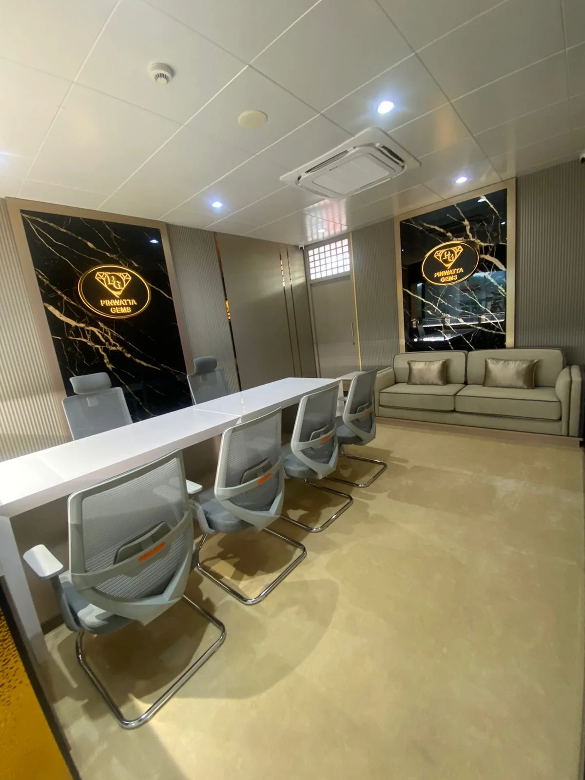 Modern conference room with white table, six gray chairs, beige sofa, black marble wall panels with gold logo reading 'Pinwatta Gems,' ceiling air conditioning unit, and soft lighting.