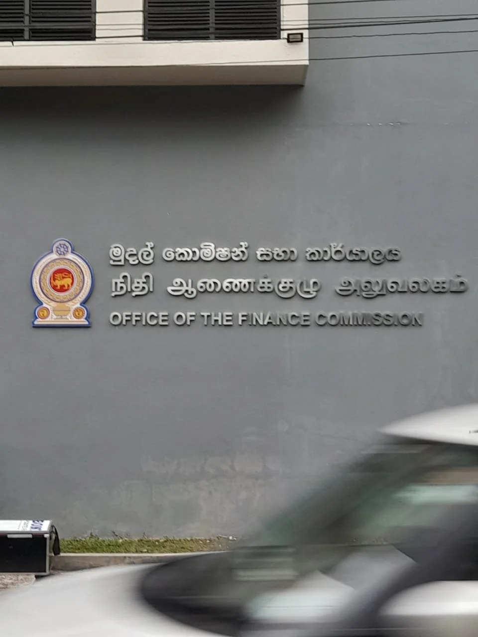 Sign for the Office of the Finance Commission with official emblem, written in Sinhala, Tamil, and English, on a gray building wall. Blurred car passing by.