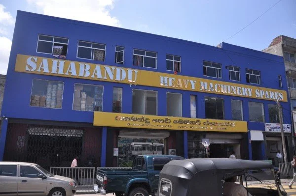 Blue commercial building with large yellow sign reading "Sahabandu Heavy Machinery Spares" and text in a local script underneath, parked vehicles in front, clear sky overhead.